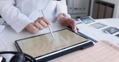 Kadın doktor dijital tablette gösterilen elektrokardiyogramı analiz ediyor. Kardiyolog klinikteki masada online ve kağıt tıbbi kayıtları karşılaştırır