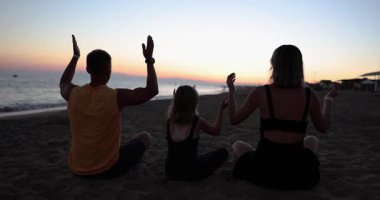 Aile bireyleri yoga sırasında yumuşak kumun üzerine oturup kollarını havaya kaldırırlar. Ebeveynler ve kızları derin bir nefes alıp temiz deniz havasının tadını çıkarıyorlar.