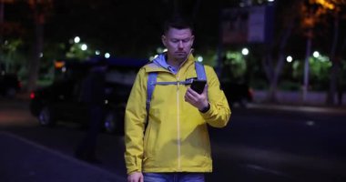 Erkek turist parlak sarı ceket giyer ve akıllı telefon kullanarak sırt çantası taşır. Gece şehir caddesinde arabalarla ve sokak ışıklarıyla dolaş.