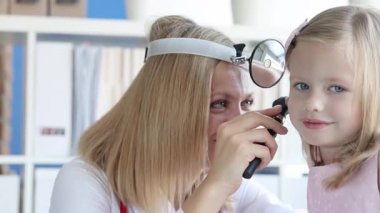 Kafa aynalı KBB doktoru muayenehanesinde otoskoplu gülümseyen küçük bir kızın kulağını inceliyor. Kadın pediatrist çocuğun kulak enfeksiyonunu kontrol ediyor