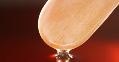Ahşap spatuladan akan balmumu damlaları sıcak kırmızımsı kahverengi arkaplanın yanında yumuşak yapışkan bir akıntı oluşturuyor. Güzellik ve tüy dökücü kavramlar için idealdir.