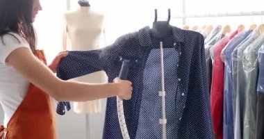 Profesyonel terzi, moda atölyesindeki buharlı gömleği askıya asar. Kadın, çalışma alanını çevreleyen raflarda ütülü giysilerle çalışıyor.
