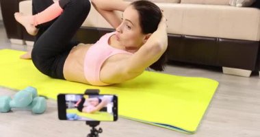 Kadın fitness antrenörü, internet üzerinden akıllı telefondan yoga minderinde uzanan ve mekik çeken spor videoları kaydediyor. Karantina sırasında sağlıklı yaşam biçimi