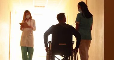 Yüz maskesi takan hemşire ve doktor tekerlekli sandalyedeki erkek hastanın yanında yürüyor. Güneşli hastane koridorundaki insanlar umut ve destekle dolu.
