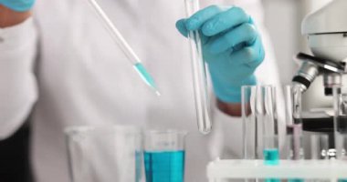 Koruyucu eldivenli laboratuar asistanı masasındaki test tüpüne mavi sıvı damlatıyor. Teknisyen, bilimsel araştırma merkezinde kimyasal reaktörle çalışıyor.