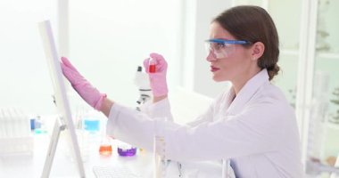 Kadın bilim adamı laboratuvar ortamındaki bilgisayarlarda test sonuçlarını karşılaştırmak için örnek şişesi tutuyor. Yoğunlaştırılmış asistan laboratuvardaki dijital verileri kontrol ediyor