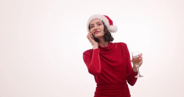 Kırmızı Noel Baba şapkalı ve elbiseli genç kadın elinde şampanya kadehiyle telefon görüşmesi yapıyor. Bayan Noel 'i beyaz arka planda ağır çekimde kutluyor.