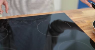 Ev hanımı, modern mutfakta indüksiyon sobasında yemek hazırlıyor. Kadın elleri tavayı evde ocağın içine yerleştiriyor.