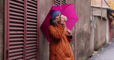 Konforlu bereli ve parlak turuncu ceketli bir kadın elinde parlak pembe şemsiyeyle kahveyi yudumluyor. Şehirde yağmurlu bir sonbahar günü huzurlu bir anda