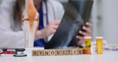 Doktor, masadaki tahta bloklarda sinovitis teşhisi konulmuş diz röntgeni görüntüsünü tutuyor ve inceliyor. Klinik ortamında eklem iltihabı teşhisi