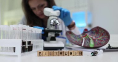 Tahta bloklar üzerinde Bilrubin kelimesi ve doktor masasındaki karaciğer modelinin kan örnekleri. Laboratuvar asistanı, sağlık hizmetlerinde mikroskop kullanarak numune üzerinde çalışıyor.