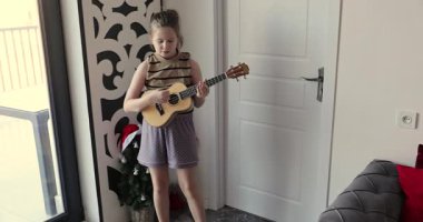 Küçük kız evdeki küçük Noel ağacının yanında ukulele çalıyor. Küçük gitarlı mutlu çocuk müzik yapıyor. Evinde keyifli anlar yaşıyor.