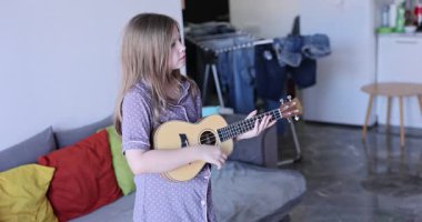 Pijamalı genç kız, oturma odasında müzik üzerine yoğunlaşarak ukulele çalıyor. Çocuk rahat ev ortamında huzurlu ve neşeli anın tadını çıkarıyor.