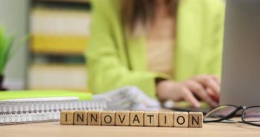 Masaya düzgünce yerleştirilmiş küçük tahta küpler kelime innovasyon oluşturur. İş kadını masada oturuyor ve belgelerin yanındaki dizüstü bilgisayarda etkili bir şekilde yazıyor.