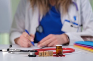 Bir sağlık uzmanı özenle notlar yazarken, masaya steteskop ve SLE ilaçları yerleştiriliyor.