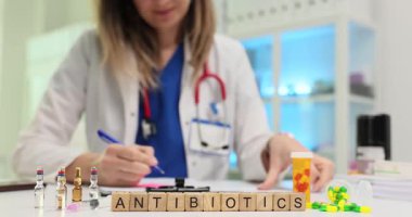 Doktor tedavi planını belgeleyecek. Doktor masasına oturur ve antibiyotik reçetesi yazar. Farklı antibiyotiklerle dolu şişeler masada duruyor.