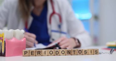 Tahta bloklara Periodontitis ve doktorun yanındaki diş modeline mesaj at. Kadın dişçi, ağız boşluğu sağlığının önemli yönlerine odaklanarak tıbbi form dolduruyor.