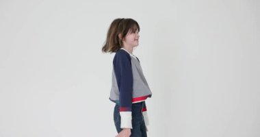 Dağınık saçlı neşeli küçük kız beyaz duvarın etrafında zıplıyor. Renkli kazak ve kot pantolon giyen oyuncu liseli kız yavaş çekimde eğleniyor.