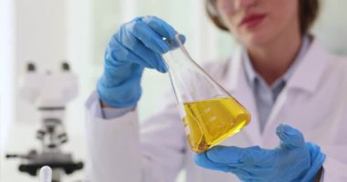 Laboratuvar önlüğü ve mavi eldivenli kadın bilim adamı Erlenmeyer şişesindeki sarı kimyasal çözeltiyi inceliyor. Klinikte mikroskop ile tıbbi araştırma