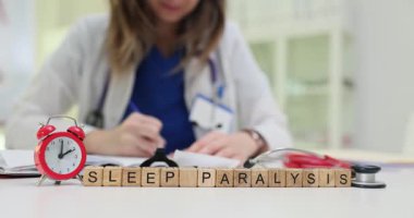 Doktor masasında, tahta küplerin uyku felci ve çalar saatin yazılı olduğu bir yazı var. Kadın hekim klinikteki beden ve akıl hastalıklarından tedavi olmayı seçiyor