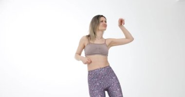 Spor elbiseli zayıf sporcu kadın beyaz stüdyo arka planına karşı neşeyle dans ediyor. Gülümseyen sarışın kadın enerjik dans hareketleri sergiliyor. Yavaş çekim