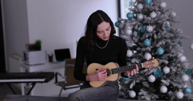 Genç bayan müzisyen, süslü Noel ağacının yanındaki koltukta ukulele çalıyor. Düşünceli kadın evdeki oturma odasında neşeli bir atmosfer yaratıyor.