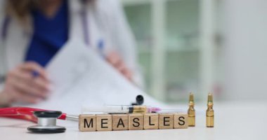 Doktor masasında kızamık ve aşı ampulleri yazan tahta küplerin yanında kağıtlarla çalışıyor. Kadın modern klinikte kızamık aşısı konsepti sağlıyor.