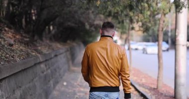 Açık kahverengi deri ceket giyen bir adam işlek caddenin yanındaki kaldırımda arabalarla yürüyor. Moda tarzı ve şehir yaşam tarzına odaklan