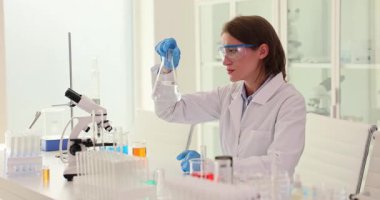 Laboratuvar önlüğü ve güvenlik gözlüğü takan kadın bilim adamı mikroskobun yanındaki Erlenmeyer şişesindeki sıvıyı analiz ediyor. Steril laboratuvarda bilimsel araştırma