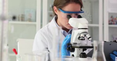 Laboratuvar önlüğü ve koruyucu gözlük takan kadın bilim adamı mikroskop kullanır ve tablet üzerinde notlar alır. Modern laboratuarda ekipmanlarla yapılan araştırmalar