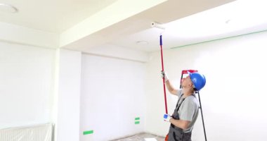 İş tulumu ve şapka giyen profesyonel bir ressam beyaz tavanı boyamak için teleskopik boya silindiri kullanır. Ev yenilenirken boş oda