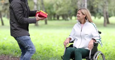 Adam tekerlekli sandalyede oturan gülümseyen kadına zarif bir hediye sunar. Erkek arkadaş, içinde sevgi ifadesi olan hediye kutusu sunar. Yavaş çekim