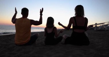 Baba ve kız gün batımında plajda yoga yapıyorlar. Yaz tatilleri sağlıklı aile alışkanlıklarını ve sağlığını farkındalık yoluyla geliştirir.