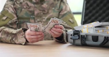 Kamuflaj üniformalı bir asker masada oturmuş yüz dolarlık banknotları sayıyor. Ofisteki adamın yanında bantlı para dolu çantaları aç.
