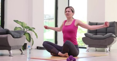 Evde yoga yapan bir kadın oturma odasında paspasın üzerinde otururken omuz ağrısı çekiyor. Kollarını esnetirken sinir sıkışması sağlıklı yaşam tarzını teşvik ediyor.