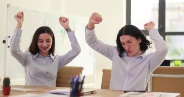 İş arkadaşları ofis masasında otururken şakacı bir şekilde dans ediyorlar. Kolları havada neşeli profesyoneller modern işyerinde iyi haberleri kutluyor.