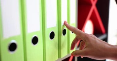 Ofis çalışanı, raftaki yeşil yüzük klasörlerine nazikçe dokunarak dosyaları inceliyor. Şirket ofisindeki önemli belgeleri düzenlemeye odaklan.