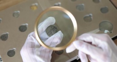 Beyaz eldiven giyen nümismatör, antik gümüş sikkeyi büyüteçle dikkatlice inceliyor. Tarihsel eserin analizini yapan kişi