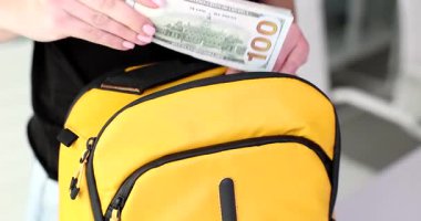 Kadın 100 dolarlık banknotları parlak sarı sırt çantasına yerleştiriyor. Yurt dışına seyahat simgesi tasarruf yatırımı ve mali planlama