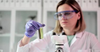 Koruyucu gözlüklü ve eldivenli kadın bilim adamı modern laboratuarda test tüpünü sarı sıvıyla analiz ediyor. Mikroskop ile bilimsel araştırma