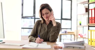 İş kadını masasında oturmuş, parlak modern ofiste telefon konuşmalarıyla meşgulken not alıyor. Doğal ışık, odaklanmış çoklu görevi artırır