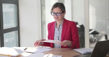 Gözlüklü ve kırmızı ceketli gülümseyen iş kadını masada otururken belgeleri gözden geçiriyor. Aydınlık ofiste dizüstü bilgisayar ve çizelgelerle çalış