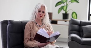 Odaklanmış, uzun gri saçlı son sınıf öğrencisi kadın evinde rahat bir şekilde kitap okumaya dalmış bir şekilde oturuyor. Hanımefendinin boş zamanları.