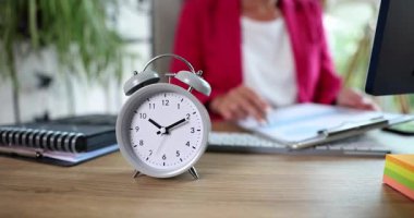 Çalar saat ahşap masada tam olarak ne zaman durduğunu gösteriyor. Parlak ceketli meşgul kadın son teslim tarihi ofis atmosferi yaratan rapor sayfalarını okuyor