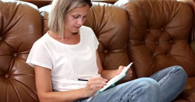 Gündelik kıyafetli sarışın kadın deri koltukta rahatça oturur ve defterine kalemle notlar yazar. Evdeki düşünceli yansıma ve yaratıcılık