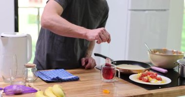 Gri tişörtlü adam mutfak tezgahında durmuş tavanın yanındaki cam bardakta içki hazırlıyor. Mutfak malzemelerinin yanında sıcak bir içecekle demlenmiş çay poşeti.