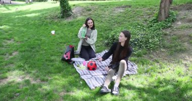 Genç kadınlar parkta çimlerin üzerinde battaniyeye oturarak rahatlar. En iyi arkadaşlar çantalardan mekikleri ve raketleri çıkarıp aktif oyunlar oynamaya hazırlanıyor.