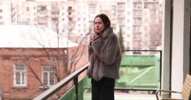 Kürk ceketli genç kadın şehir manzarasına karşı balkonda kahve içiyor. Bayan etrafına bakıyor ve kış sabahı düşüncelere dalmış.