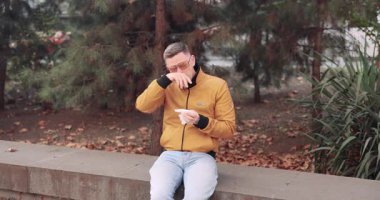 Sarı ceketli bir adam soğuk algınlığı belirtisi olarak mendil kullanarak korkuluğa oturur. Sonbaharda ağaçlar ve çalılarla kaplı parkta meydana gelen soğuğa bağlı hastalık.