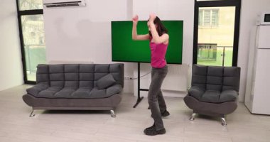 Kadın evde kromakey TV ile müzik videosu izleyerek neşe içinde dans ediyor. Gülümseyen bayan elini kaldırır ve oturma odasında eğlenerek döner.